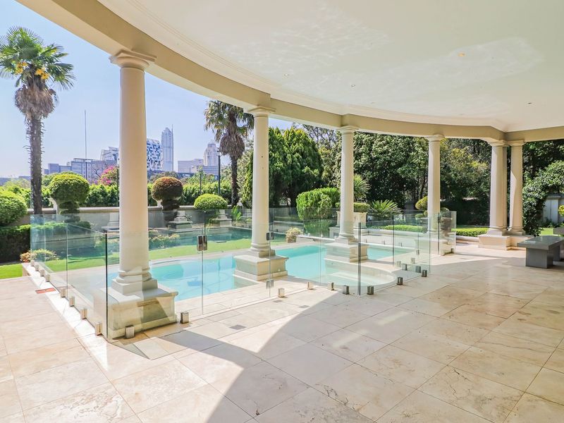 A spacious covered patio featuring stone flooring, elegant columns, and a glass-fenced swimming pool with a view of the city skyline.