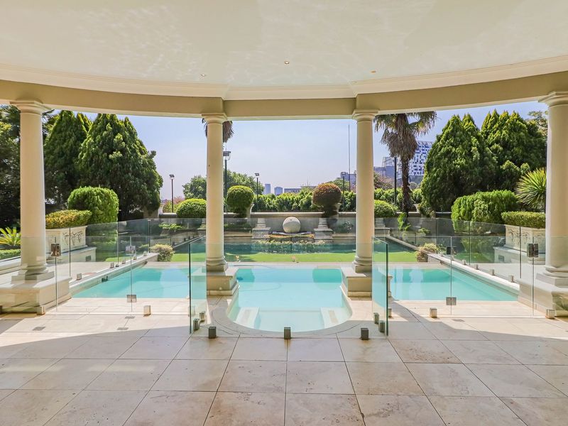 A luxurious covered patio overlooking a pristine swimming pool with glass balustrades, classical columns, and manicured garden surroundings.