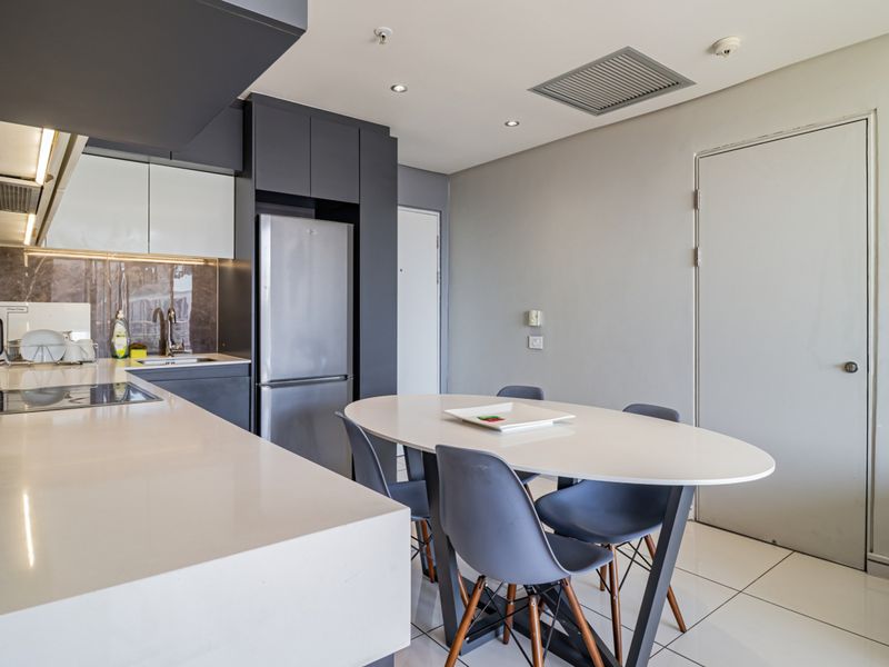 Modern open-plan kitchen and dining area featuring sleek dark cabinetry, white quartz countertops, integrated appliances, and polished tiled flooring.