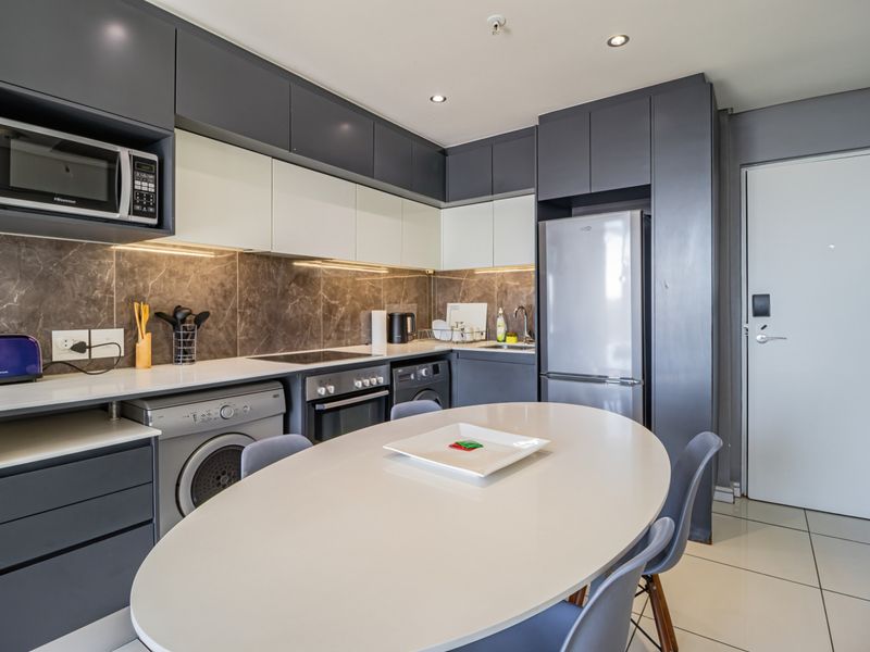 Modern open-plan kitchen featuring sleek grey and white cabinetry, stone countertops, integrated appliances, and tiled flooring in a well-lit, contemporary apartment setting.