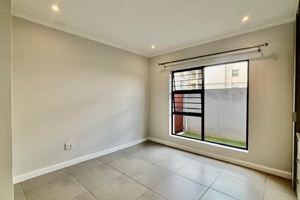 A bright, empty bedroom featuring neutral tiled flooring, recessed downlighting, and a large aluminium-framed window with a curtain rod.