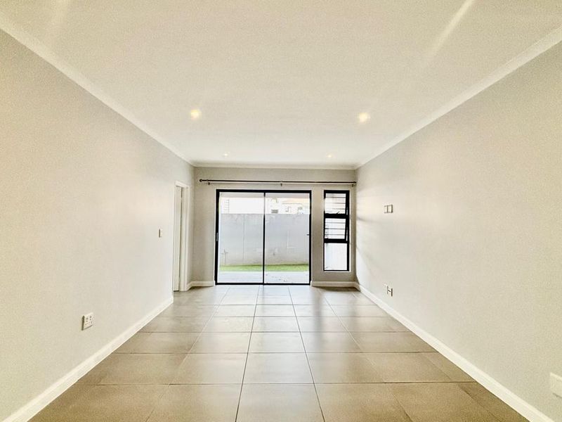 A bright, empty living room featuring neutral tiled flooring, recessed lighting, and a sliding glass door providing access to the outdoor area.