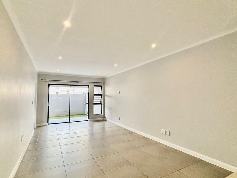 A bright, empty living room featuring neutral tiled flooring, recessed downlighting, and a sliding glass door providing access to the outdoor area.