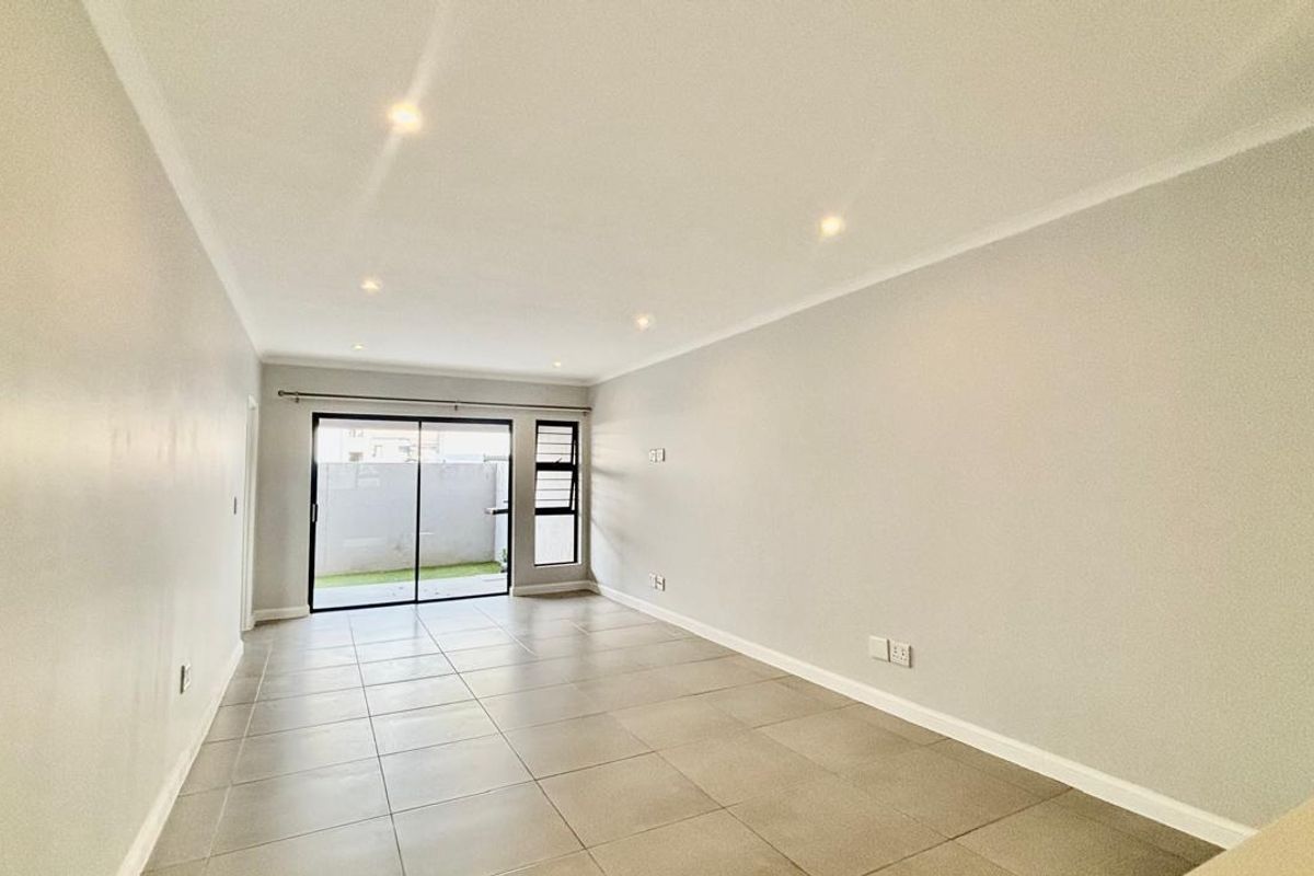 A bright, empty living room featuring neutral tiled flooring, recessed downlighting, and a sliding glass door providing access to the outdoor area.