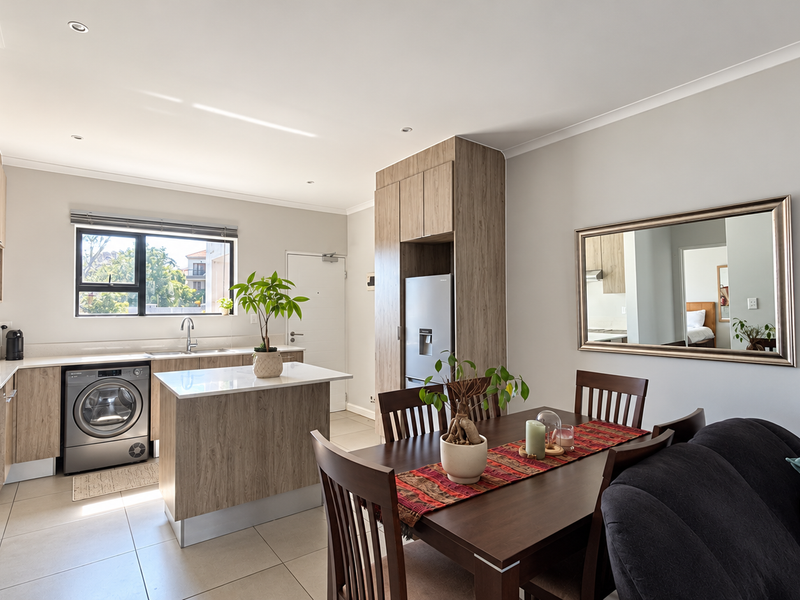 A modern open-plan living and kitchen area featuring wood-finish cabinetry, stone countertops, integrated laundry, and neutral tiled flooring throughout.