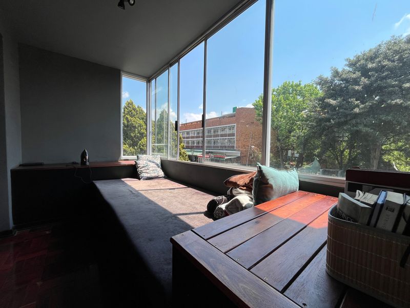 A sunlit enclosed balcony featuring large glass windows, parquet flooring, and a built-in wooden bench, offering pleasant urban views.