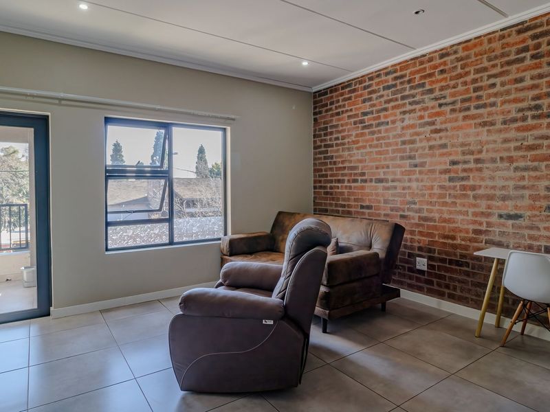 A bright living area featuring a striking exposed brick feature wall, neutral tiled flooring, and direct access to an outdoor balcony through a glass door.