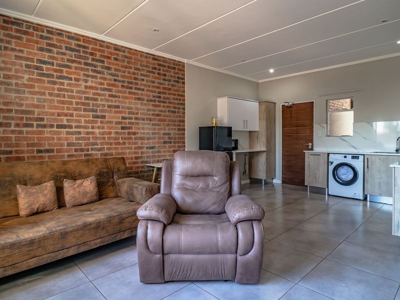 An open-plan living and kitchen area featuring a striking exposed brick wall, modern tiled flooring, and a functional kitchen with integrated cabinetry.