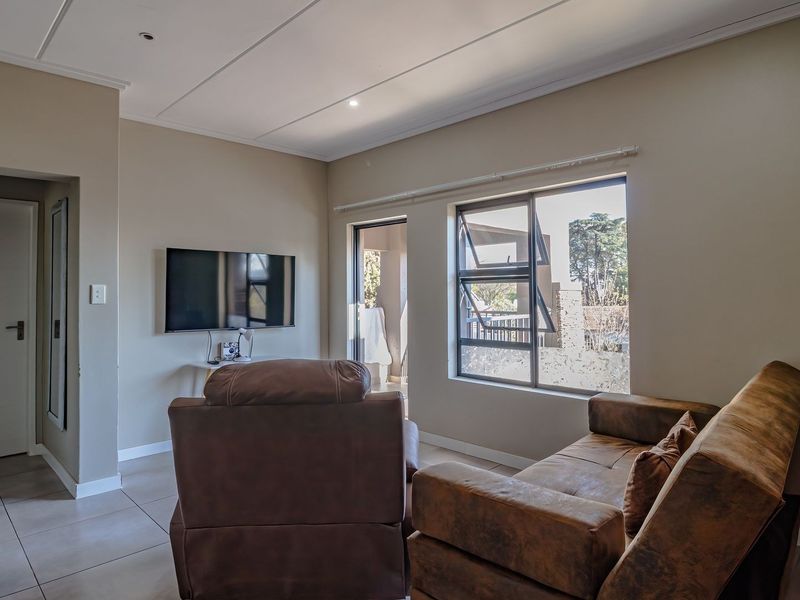 A well-lit living room featuring neutral wall tones, tiled flooring, and large aluminium windows providing natural light and access to an outdoor area.