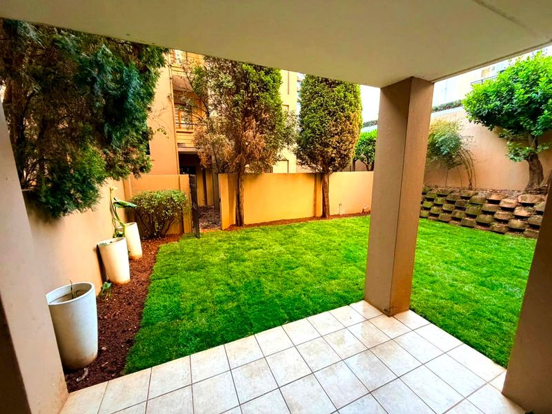 A covered patio area featuring tiled flooring, overlooking a private garden with a well-maintained lawn and retaining wall.
