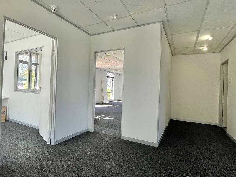 A bright, open-plan office space featuring neutral partition walls, suspended ceilings with recessed lighting, and dark grey carpeting throughout.
