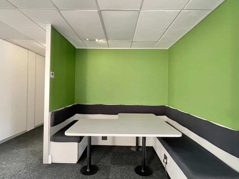 A modern office breakout area featuring built-in booth seating with integrated table, carpeted flooring, and a suspended ceiling.