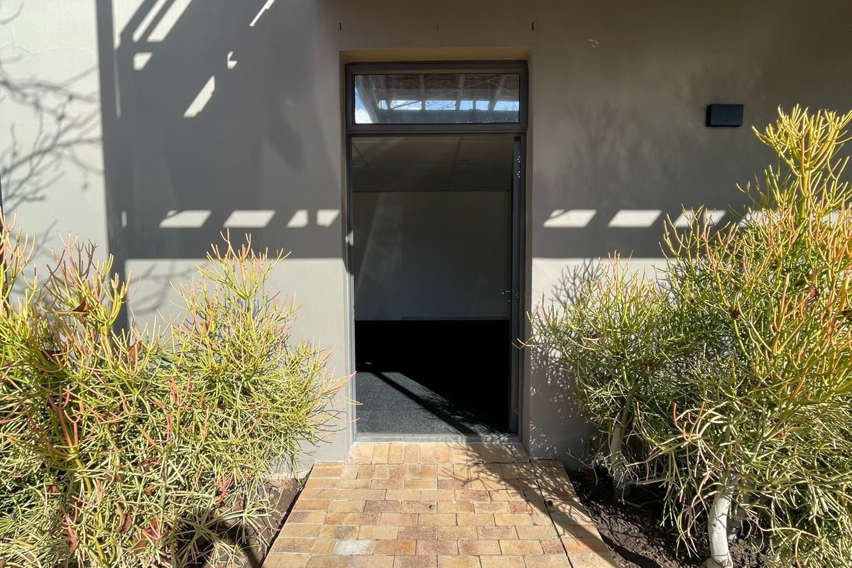 Exterior view of an office entrance featuring a brick paved walkway, metal door frame with transom window, and exterior wall lighting.