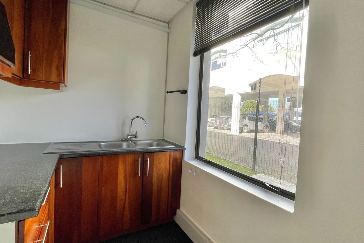 A compact office kitchenette featuring wood-finish cabinetry, a granite countertop, and a stainless steel sink with a view through a large window.