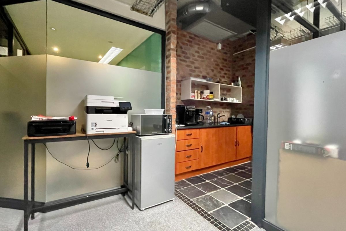 A compact office kitchenette featuring exposed brickwork, dark stone flooring, built-in wooden cabinetry, and a glass partition wall.