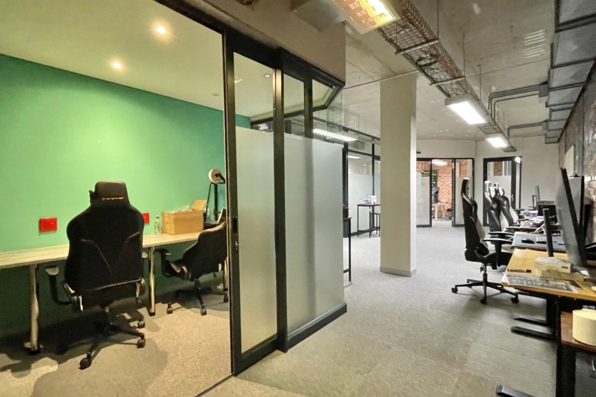 Modern open-plan office space featuring glass-partitioned meeting rooms, exposed industrial ceilings with cable management, and a mix of brick and painted wall finishes.