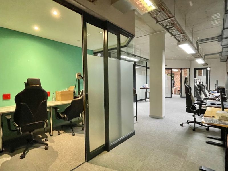 Modern open-plan office space featuring glass-partitioned meeting rooms, exposed industrial ceilings with cable management, and a mix of brick and painted wall finishes.