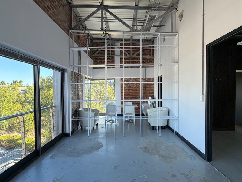 Modern industrial-style office space featuring exposed brick walls, steel roof trusses, concrete flooring, and large sliding glass doors leading to a balcony.