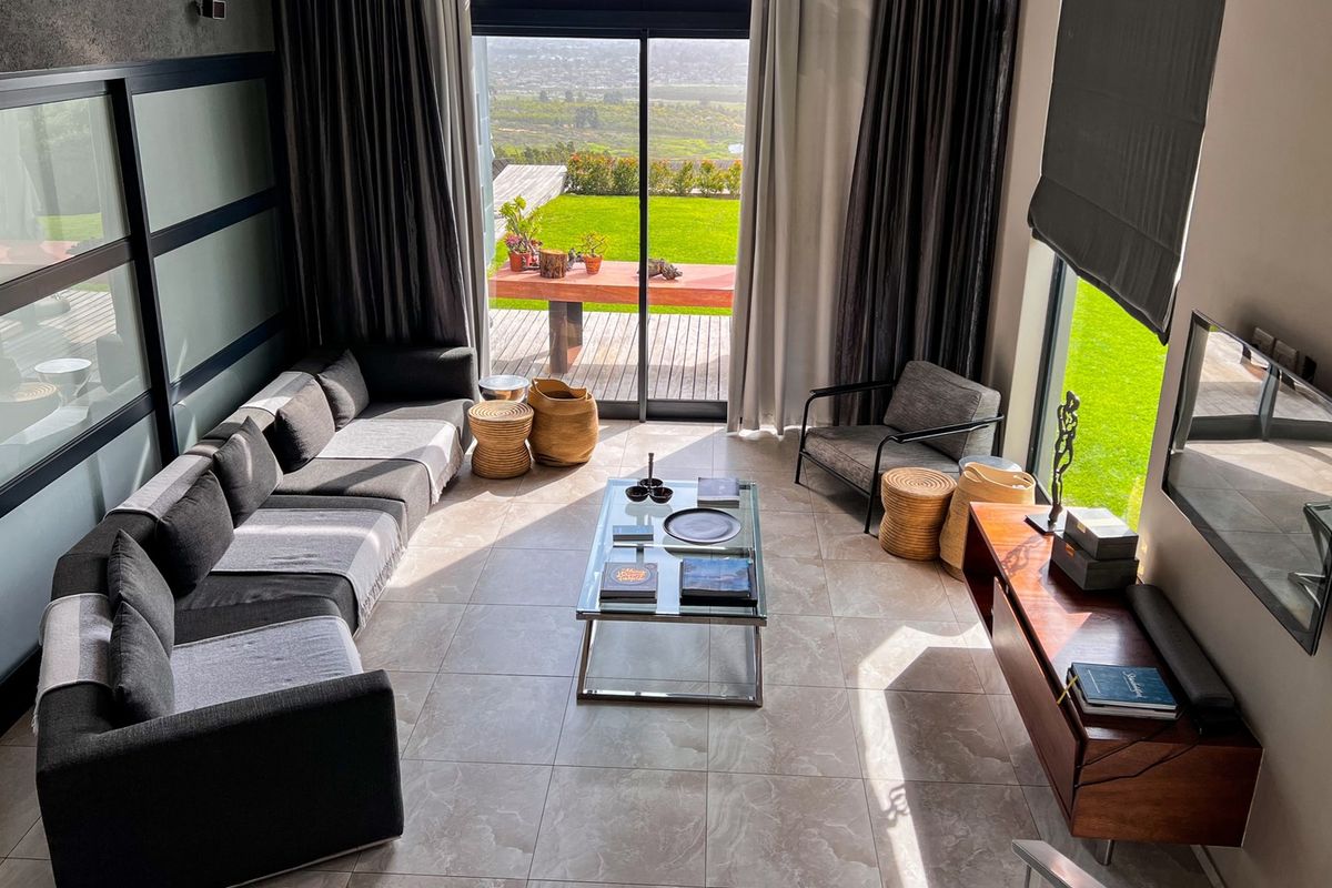 A modern, double-volume living room featuring large format floor tiles, expansive floor-to-ceiling glass doors, and abundant natural light with views of the garden.