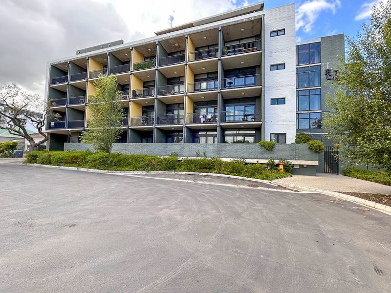 Modern multi-story apartment building featuring a concrete facade, private balconies with metal railings, and a paved driveway entrance.