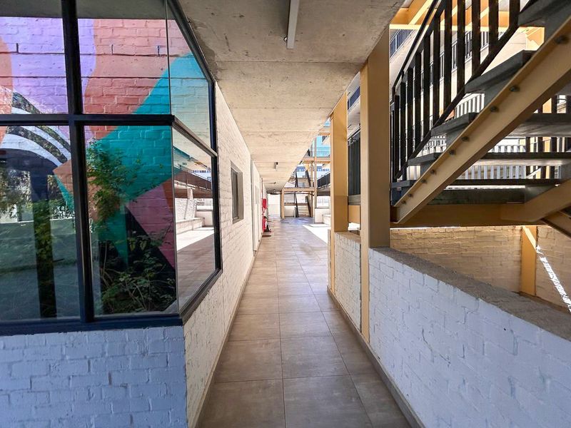 An outdoor apartment complex hallway featuring tiled flooring, exposed concrete ceilings, and a metal staircase with painted brick walls.