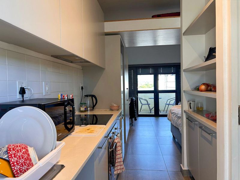 Compact galley-style kitchen featuring modern cabinetry, integrated appliances, and a tiled backsplash, leading directly into an open-plan living area with balcony access.
