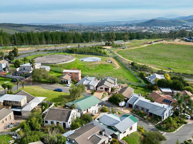 An aerial view of a residential neighborhood featuring multiple houses, paved streets, and surrounding green landscape with a water reservoir in the distance.