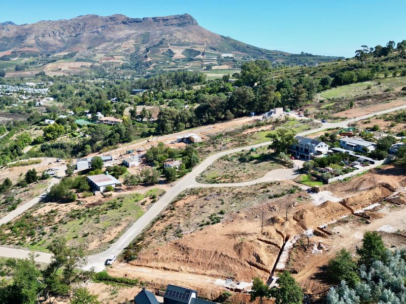 Aerial view of a residential development site featuring sloped terrain, paved access roads, and ongoing earthworks with scenic mountain views in the background.