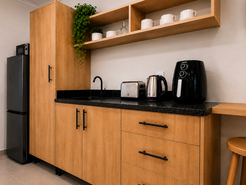 A modern, compact kitchen featuring light wood cabinetry, black hardware, a dark stone-look countertop, and integrated shelving.