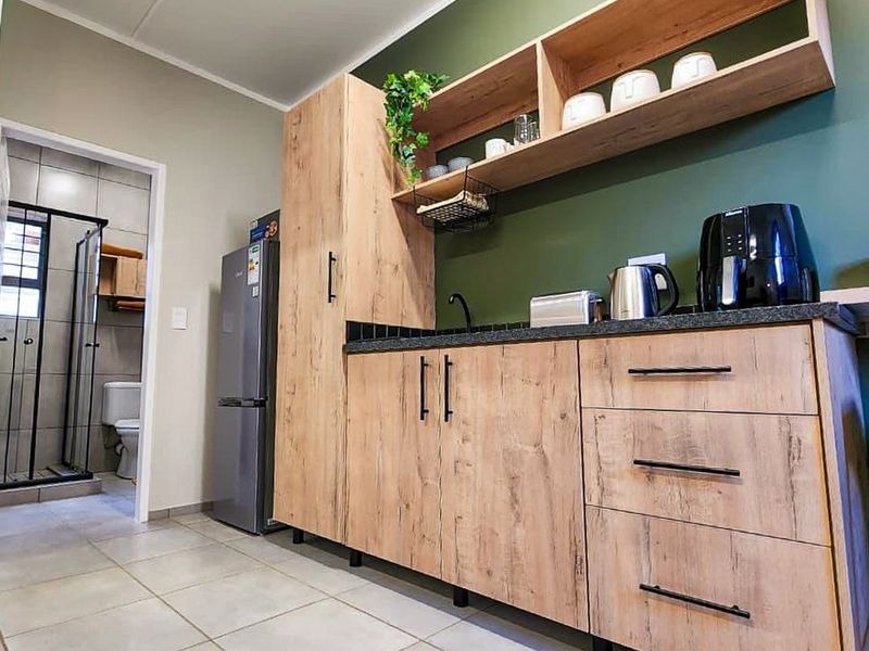 A modern kitchenette featuring wood-look cabinetry, dark countertops, and open shelving, with an adjacent view into a tiled bathroom.