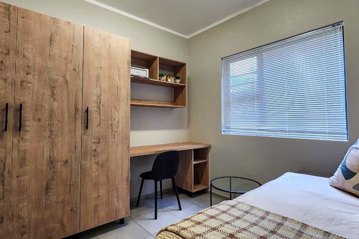 A well-maintained bedroom featuring built-in wooden cabinetry, a study desk with shelving, tiled floors, and venetian blinds for privacy.