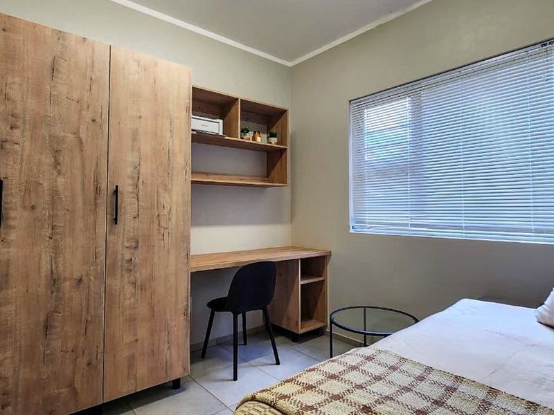 A well-maintained bedroom featuring built-in wooden cabinetry, a study desk with shelving, tiled floors, and venetian blinds for privacy.