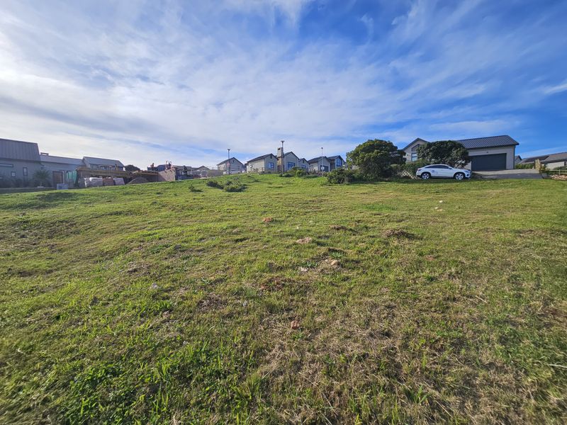 A vacant residential plot featuring a gentle slope and established grass, situated within a developed neighborhood setting.