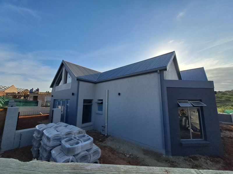 A rear view of a modern house under construction, featuring a pitched corrugated metal roof, plastered walls, and aluminium window frames.