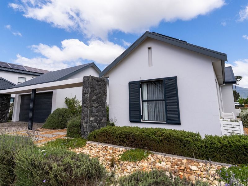A modern single-story house exterior featuring a stone-clad pillar, dark metal roofing, solar panels, and well-maintained landscaped gardens with a paved driveway.