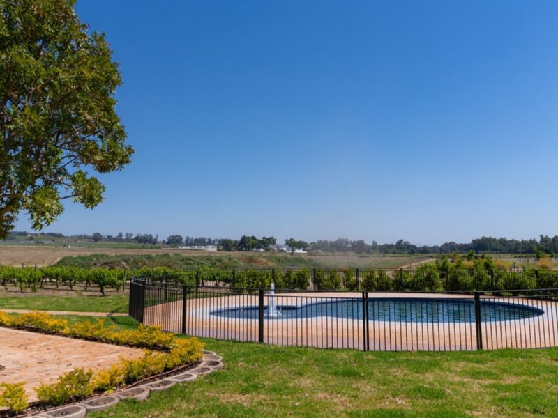 A well-maintained swimming pool area featuring a central water fountain, secure perimeter fencing, and paved surround, set against a scenic rural landscape.