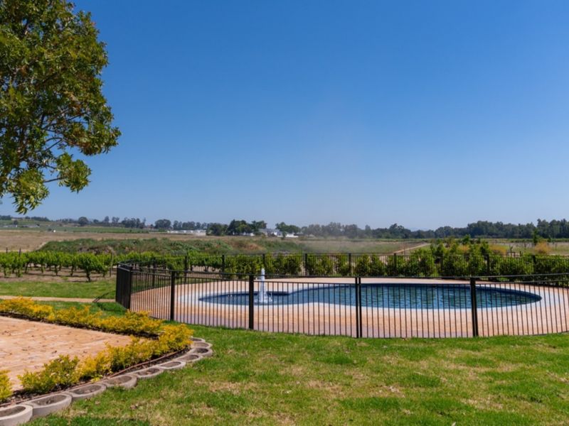 A well-maintained circular swimming pool with a water fountain feature, enclosed by metal fencing and surrounded by landscaped garden areas.