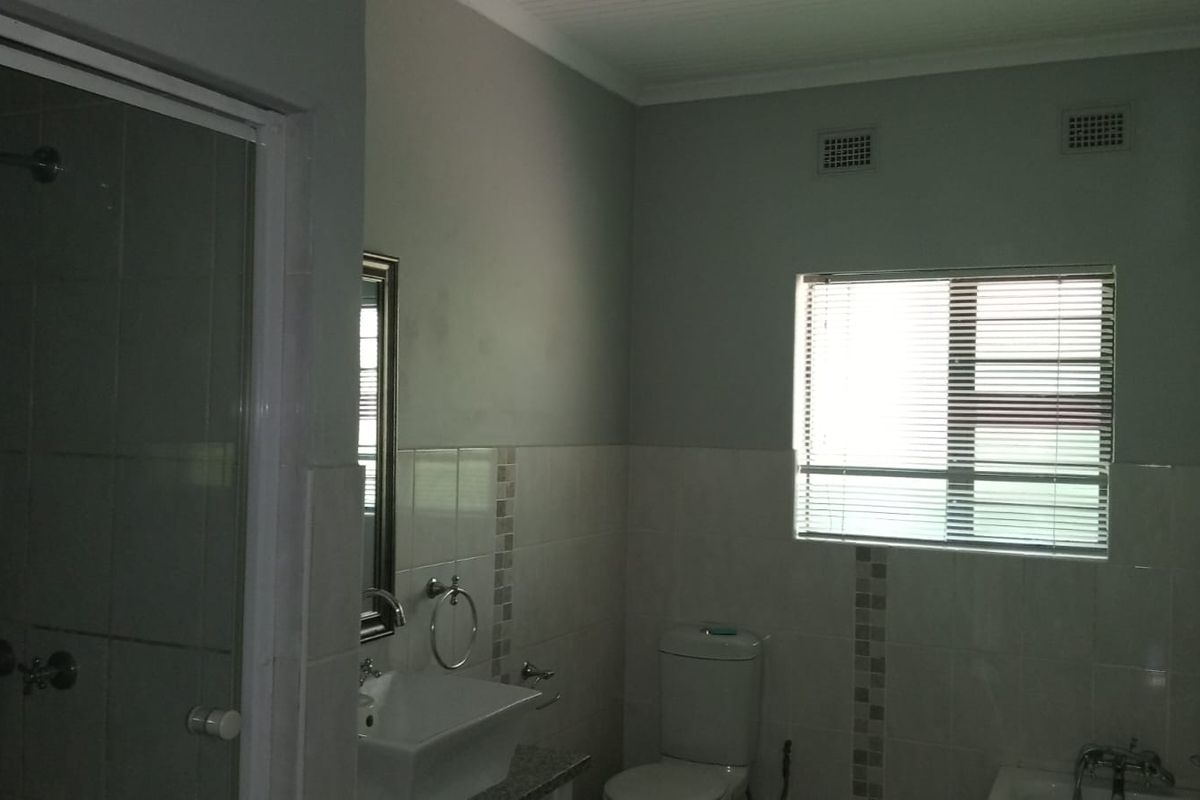 A functional bathroom featuring a glass-enclosed shower, a wall-mounted basin, a toilet, and a window with fitted blinds.