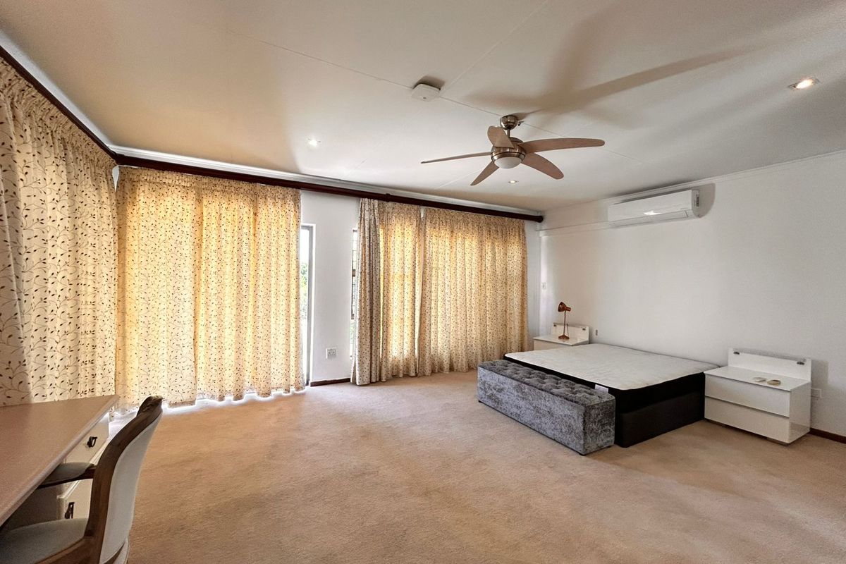 Spacious bedroom featuring neutral carpeting, a ceiling fan, and a wall-mounted air conditioning unit, with large windows providing ample natural light.