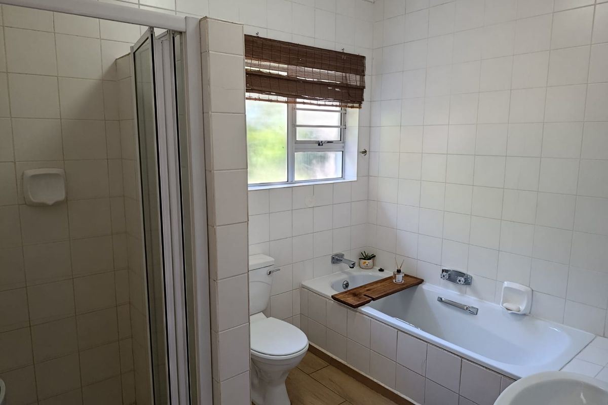 A functional bathroom featuring a built-in bathtub, separate shower enclosure, and toilet, finished with white wall tiles and wood-look floor tiles.