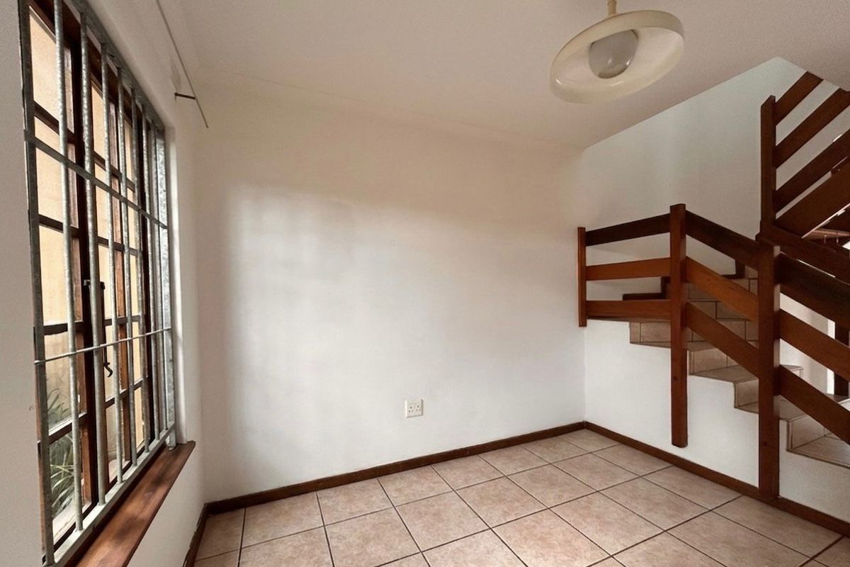 An empty living area featuring tiled flooring, a large window with security bars, and a wooden staircase leading to the upper level.