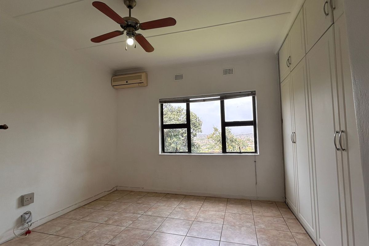A bright, empty bedroom featuring tiled flooring, built-in floor-to-ceiling wardrobes, a ceiling fan, and a wall-mounted air conditioning unit.