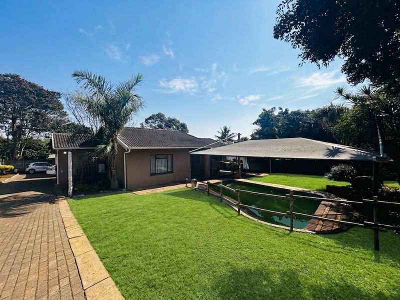 Rear exterior view of the property featuring a swimming pool with shade sail, paved driveway, and established garden area.