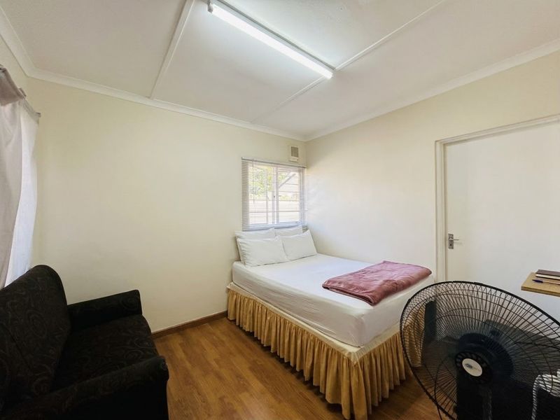 A functional bedroom featuring laminate flooring, neutral walls, and a window with venetian blinds, providing a simple and practical living space.