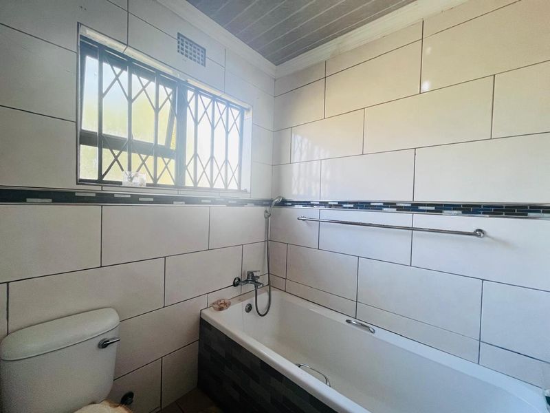 A clean, tiled bathroom featuring a bathtub with a shower mixer, a toilet, and a window fitted with security bars.