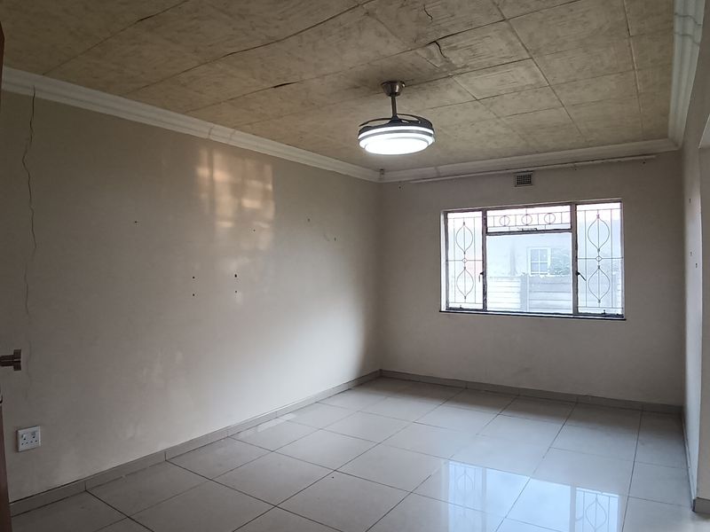 An empty living room featuring tiled flooring, a ceiling fan, and security bars on the window, showing signs of wear on the ceiling and walls.