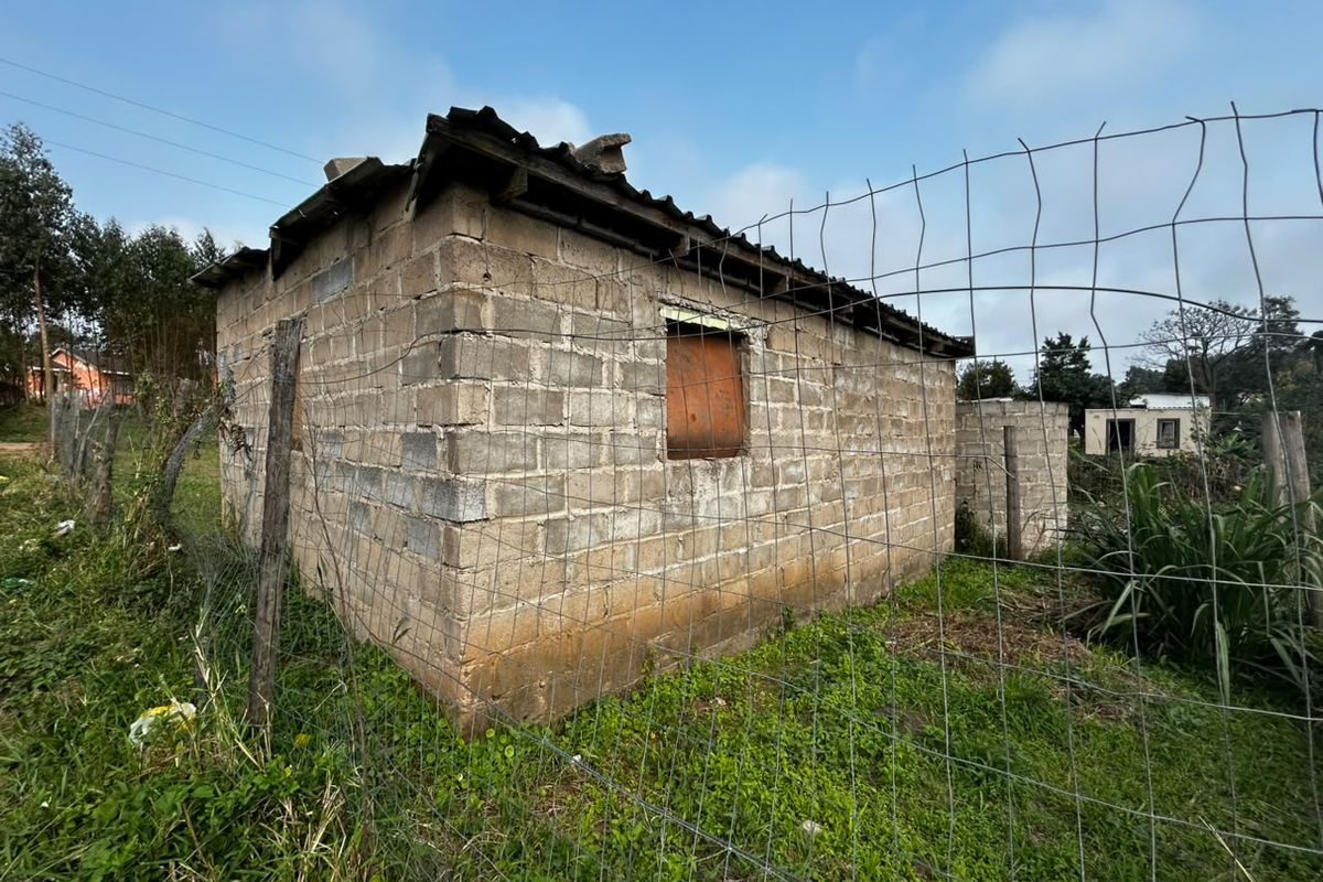 A small, unfinished concrete block structure with a corrugated metal roof, situated on a plot enclosed by wire mesh fencing.