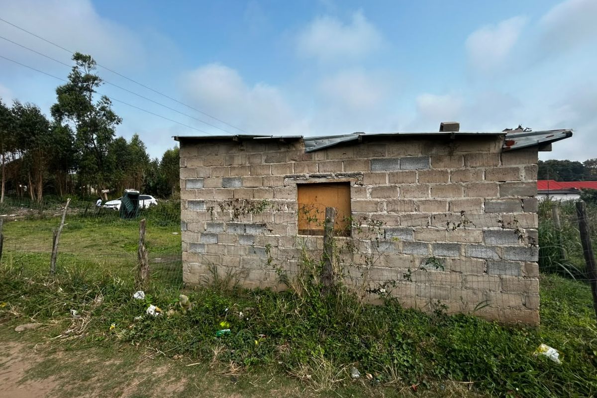 A view of a small, basic concrete block structure with a corrugated metal roof, situated on a plot of land with wire fencing.