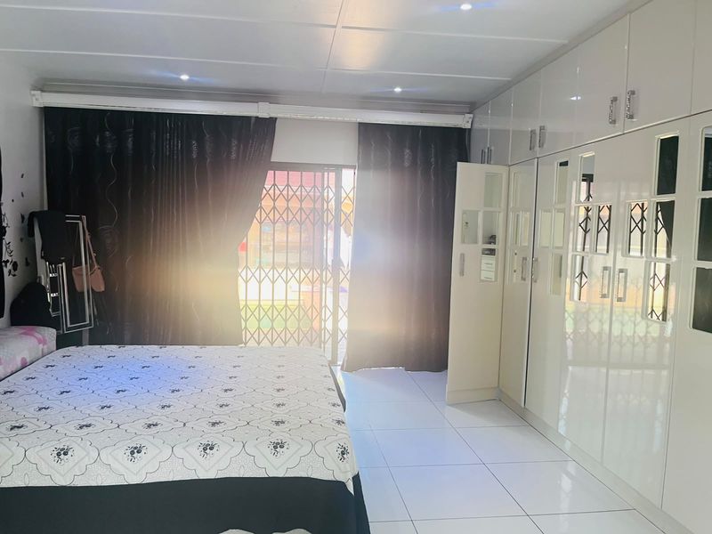 Spacious bedroom featuring glossy built-in cupboards, white tiled flooring, recessed lighting, and a sliding door with security gate leading to the exterior.