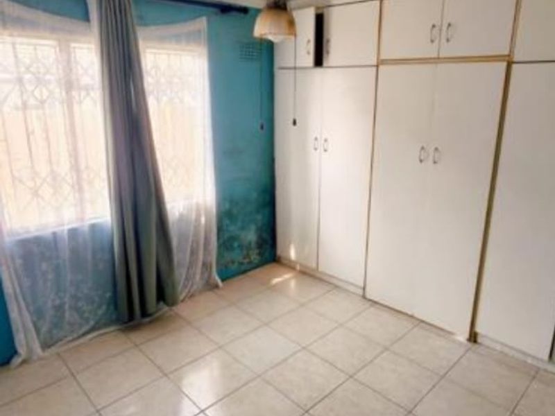 A bedroom featuring built-in cupboards, tiled flooring, and a ceiling fan. The room shows signs of wall damp and requires maintenance.
